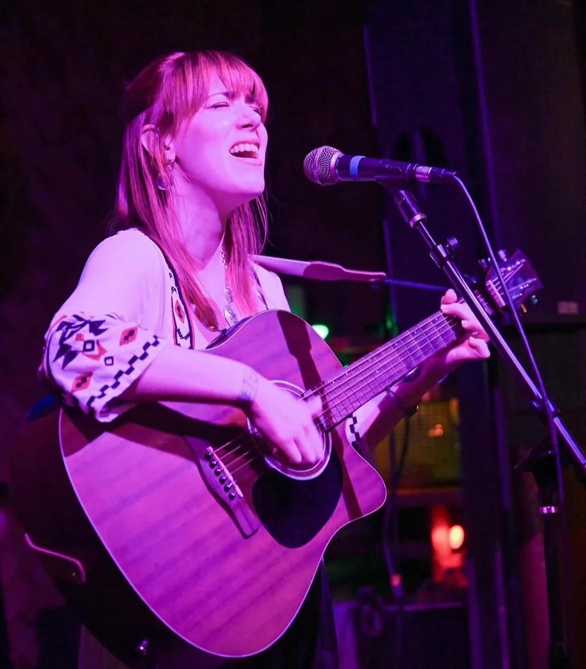 Bree Cole singing and playing acoustic guitar in magenta stage light