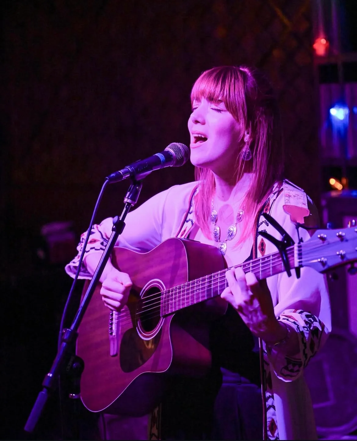 Bree Cole singing and playing acoustic guitar under purple stage lights